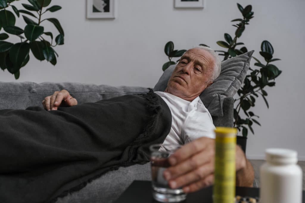 A sick elderly man lying down on sofa while holding a drinking glass